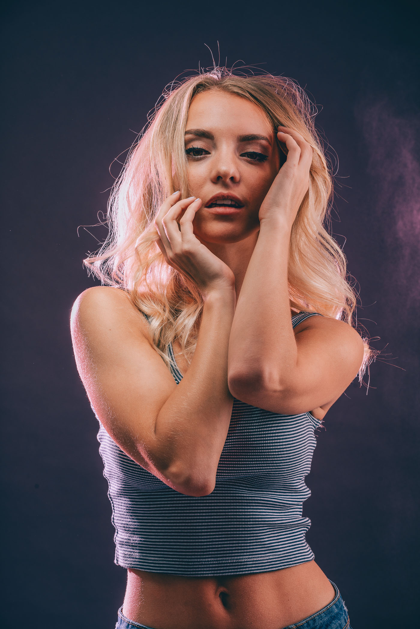 Studio portrait photography of model Anna Hrehlevych in a striped crop top against a navy and pink background with her hands by her face
