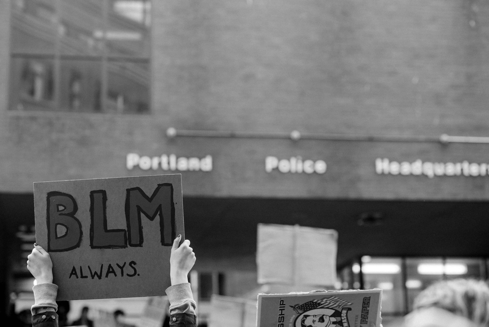 black and white photo of hands holding up a protest sign saying "BLM always" in front of Portland, Maine police station