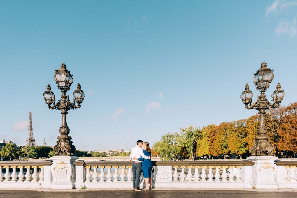 engagement photography session in paris on the pont alexandre iii bridge, wide shot with the couple kissing in the center and the eiffel tower in the distance on a sunny day