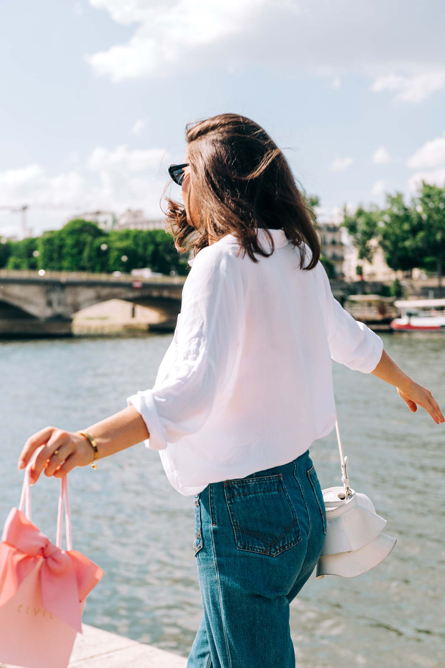 windswept lifestyle image of model on the banks of the seine wearing elvie jewelry and carrying the brand's small shopping bag as she walks