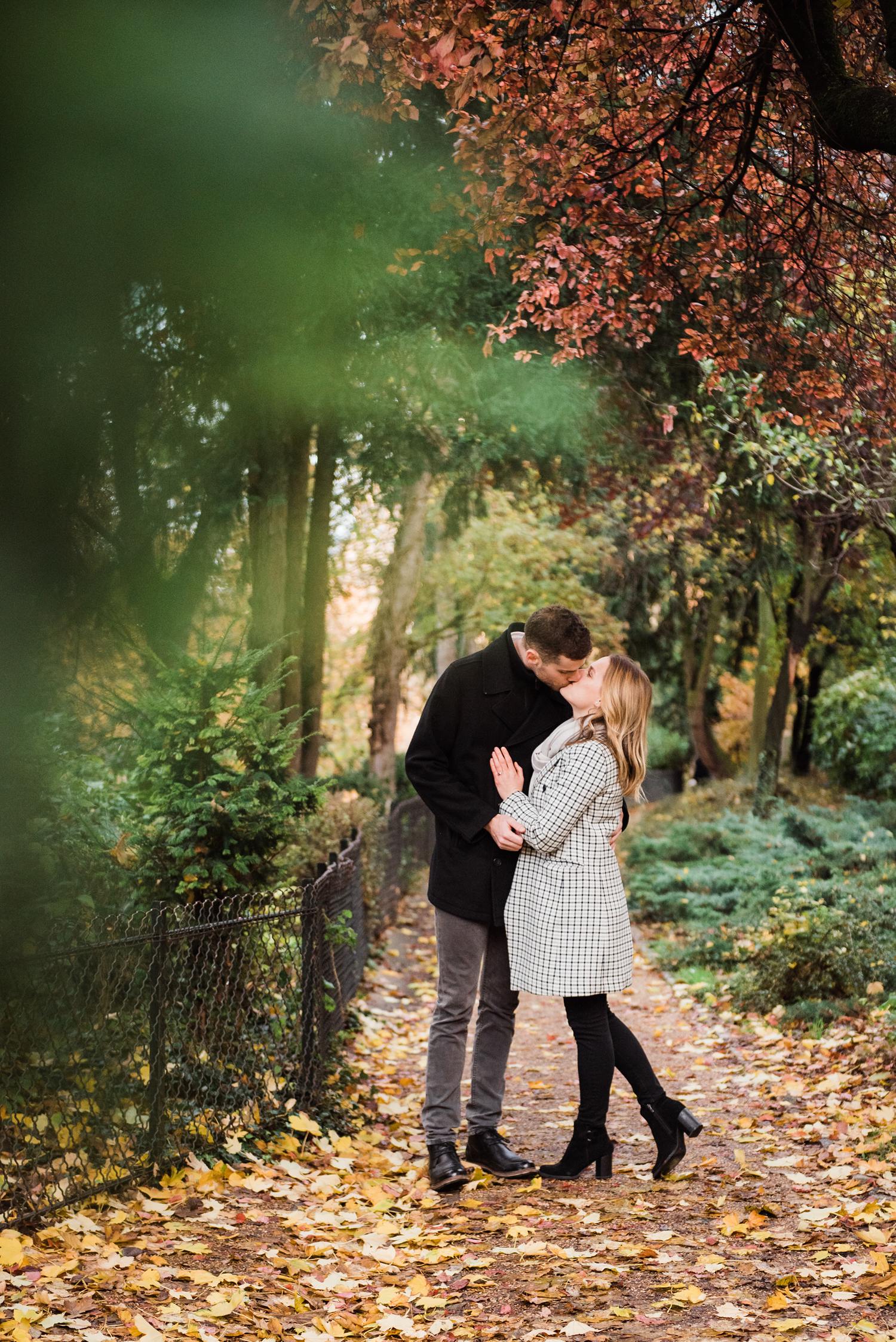 photo of couple kissing on a garden pathway at the beginning of fall with trees just starting to turn: part of an engagement photography session in Paris