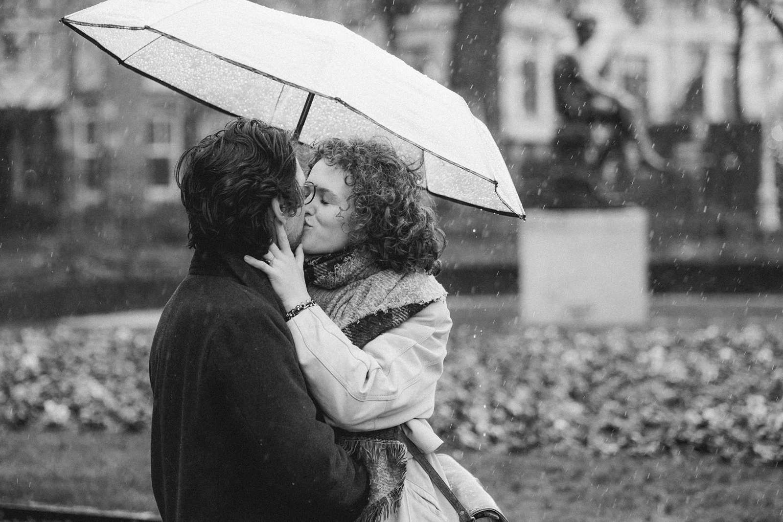 A couple shares a kiss under an umbrella, with the blurred park background adding a dreamlike touch to their intimate moment during a couple’s photoshoot. Captured by an Amsterdam photographer, this scene embodies romance in the rain.