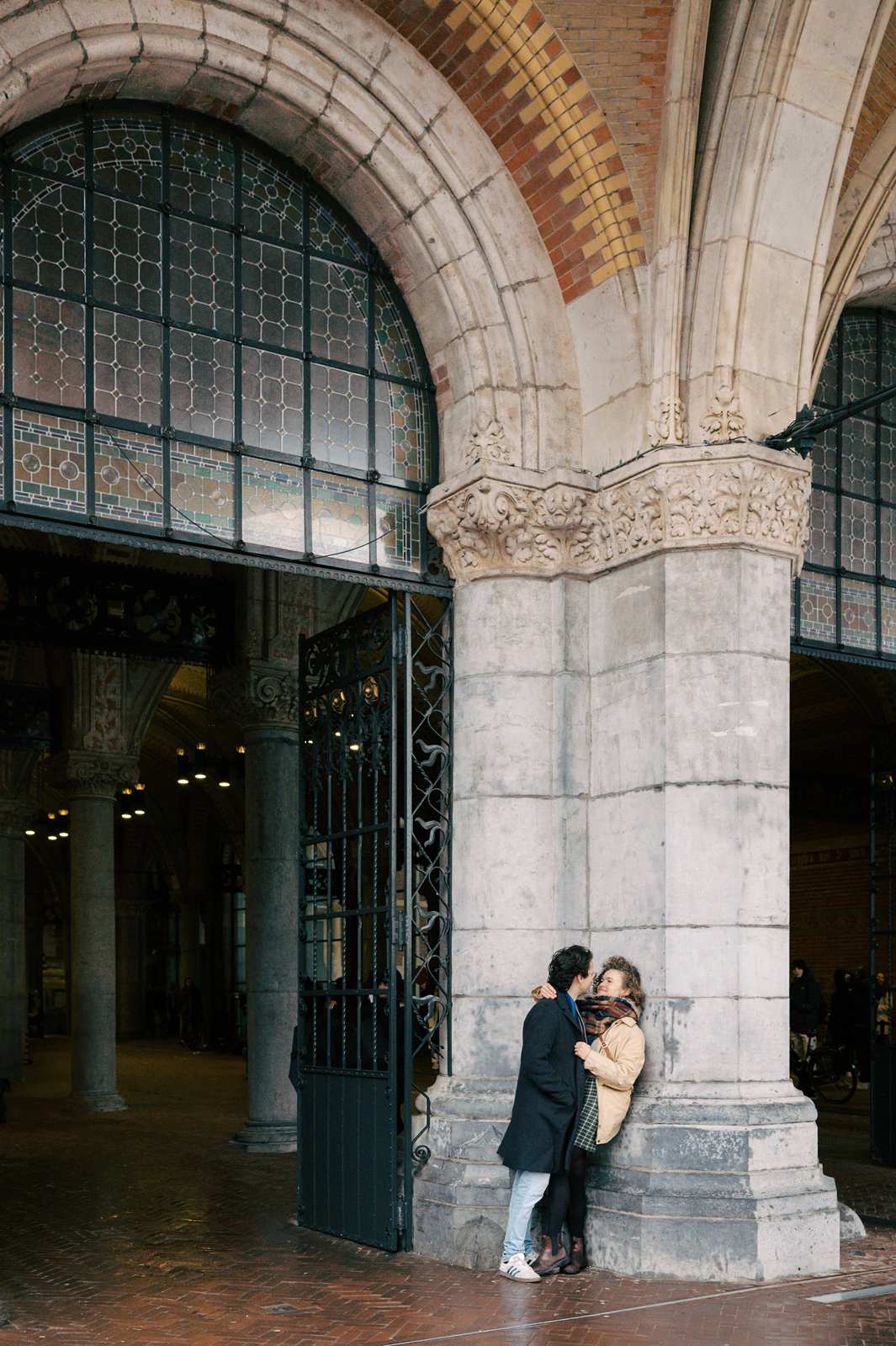 A couple leans against a stone archway with decorative metal gates at the Rijksmuseum, with their arms around each other during a romantic photoshoot in Amsterdam.