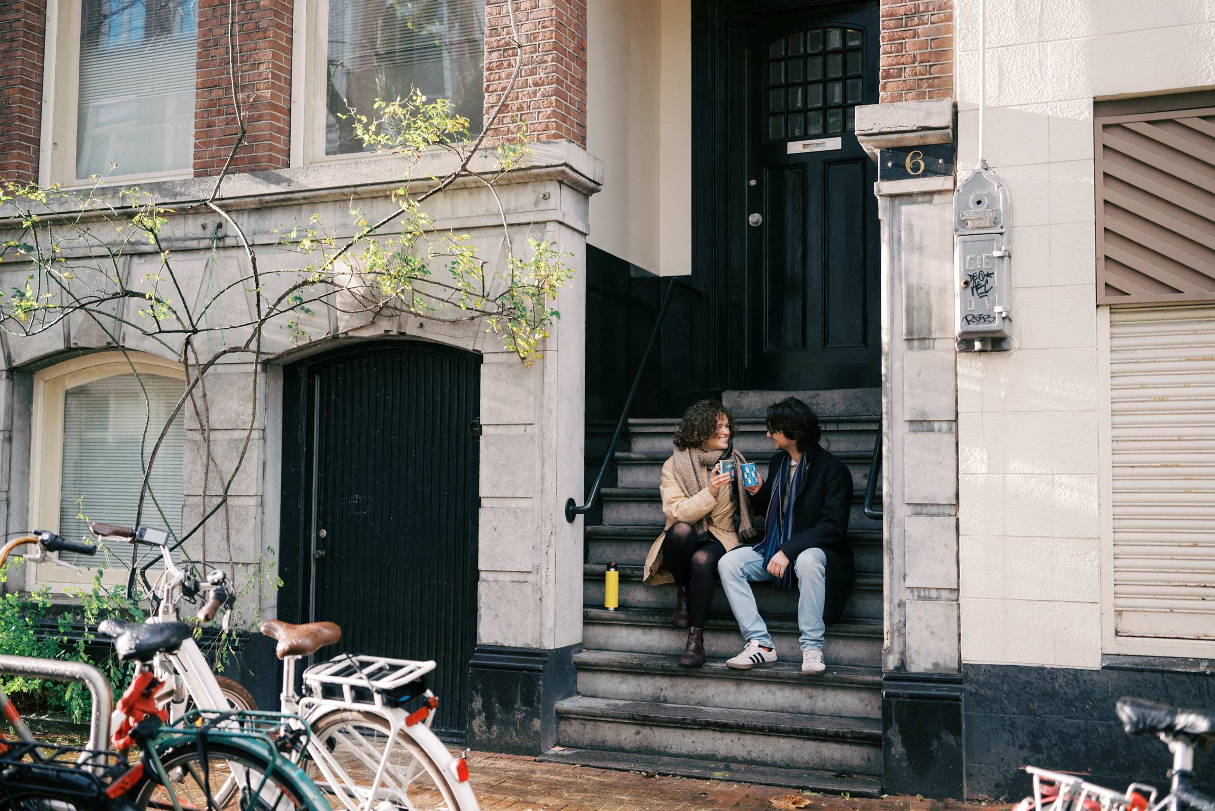 Two people sitting on stairs outside a brick building in de Pijp, talking and smiling while sharing homemade coffee. A meaningful moment captured by an Amsterdam photographer.