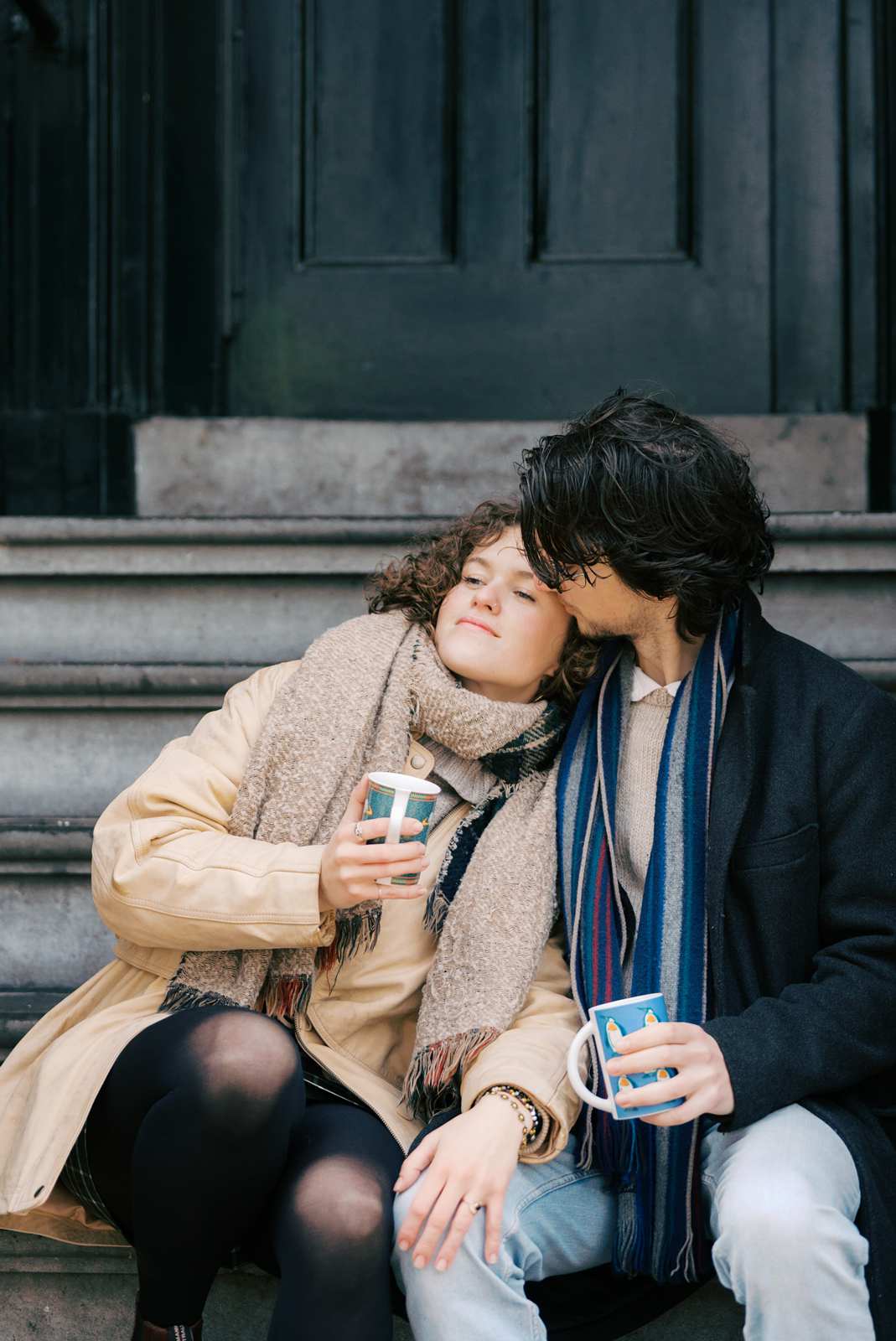 A couple sits on a stoop in Amsterdam's de Pijp neighborhood, wrapped in scarves and sipping from mugs. One person looks off into the distance, with her hand showing an engagement ring resting on her partner's knee, he kisses her on the temple. Image by professional photographer in Amsterdam Paige Gribb