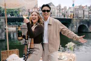 Two people smiling at a picnic by a canal, holding a wine glass and champagne bottle, celebrate during a surprise proposal, with a bridge in the background—perfect for memorable proposal photography.