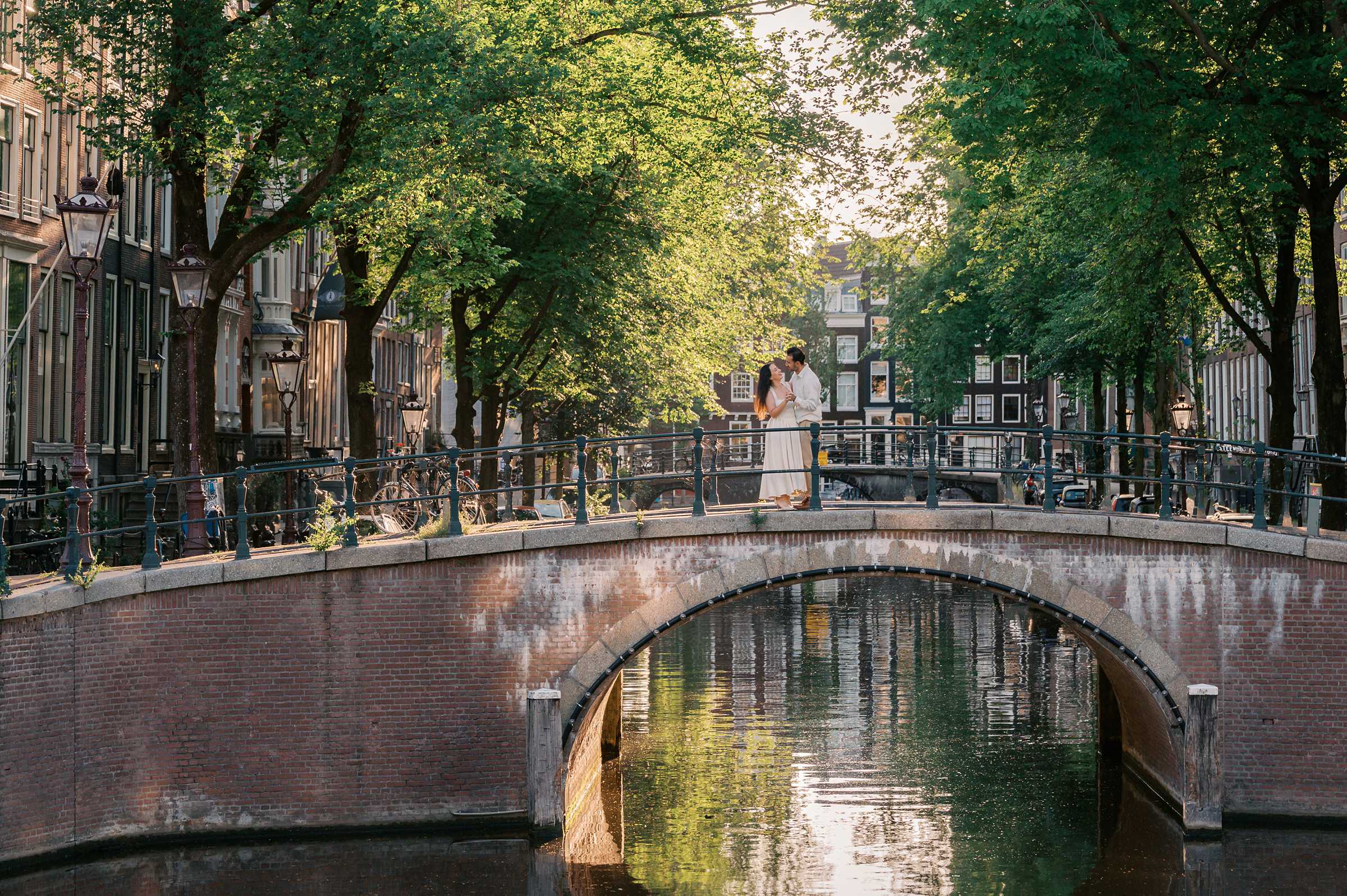 A couple cuddles together on an arched bridge over a canal in Amsterdam as part of an anniversary photo shoot in the summer