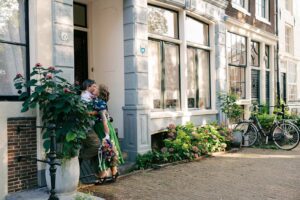 A couple kisses in the doorway of a classic Amsterdam building, holding flowers, with bicycles and plants nearby, during their sweet anniversary photo session