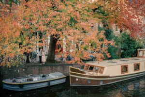 A woman stands by autumn trees on a canal at golden hour, next to two boats in Amsterdam.